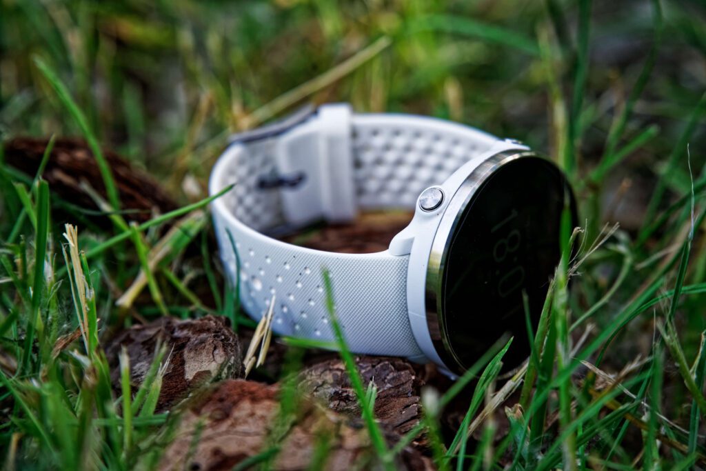 A white smartwatch with a perforated band lying on grass and dried leaves. The watch has a dark circular display and silver bezel, captured in a shallow depth of field photo.
